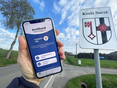 Auf einem Smartphone ist die Funkloch-App zu sehen und im Hintergrund das Kreis Soest Wappenschild. Foto: Birgit Kalle/ Kreis Soest